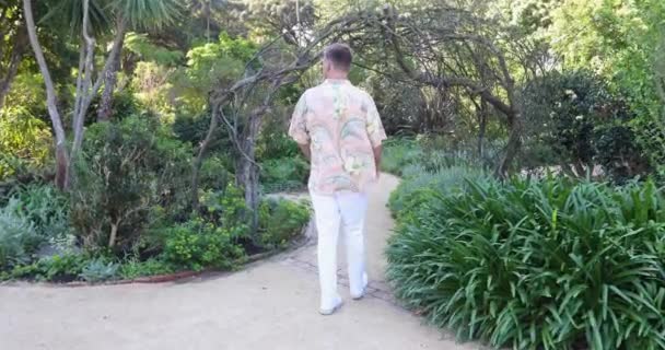 Νεαρός Άνδρας Χαβανέζικο Πουκάμισο Περπατά Μέσα Από Ξύλινη Αψίδα Garden — Αρχείο Βίντεο