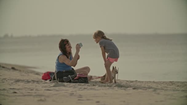 幸せな若い女性は 携帯電話で笑顔の小さな娘の写真を撮る チワワ犬と砂浜に座っている家族 — ストック動画