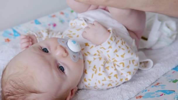 Szőke hajú, kék szemű baba bámul a kamerába, cumit szopogatva, miközben az anyja tisztítja a fenekét.