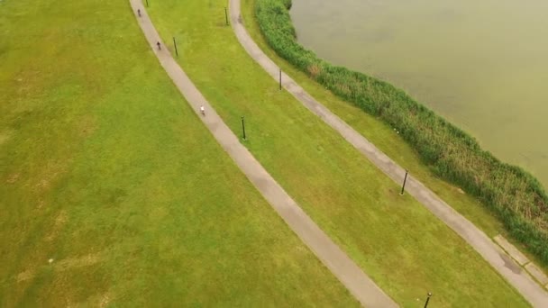 3人乗りの舗装された道を鳥の目で見ることができます 池のある緑豊かな公園では曇りの日です 自転車を追いながら下に傾いたドローンカメラ — ストック動画