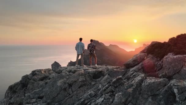 壮大な崖の上に立っている2人の男と背景に昇る太陽の空中軌道ショット — ストック動画