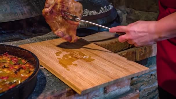 木製のオーブンの横に豚肉の肉をスライスした女性と豆と鋳鉄鍋 — ストック動画