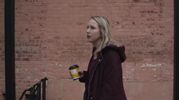 junge blonde Frau lächelt und geht tagsüber mit einem Kaffee in der Hand die Straße entlang
