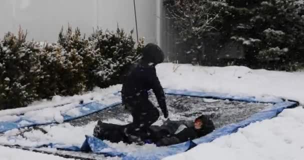 dva kluci hrající a skákající na trampolíně ve sněhových šatech během vánice, jeden dělá salto