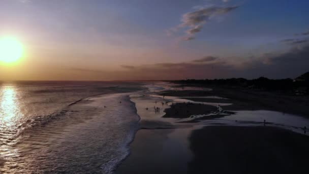 Neuvěřitelný letecký záběr barevného západu slunce na písečné pláži, lidé si hrají na pláži letící bokem k oceánu od pláže. Lidé, mávají otevřeným oceánem a štěstím. Canggu, Bali.