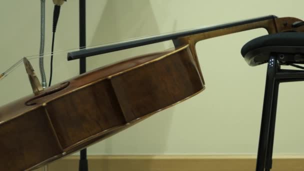 A violoncello is resting on the chair inside a recording studio. Sliding shot from the bottom of the cello and the microphone until the top and the headphones.