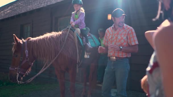 1人の小さな女の子が馬の上に座っているので ハンドラは子供たちの指示のグループを与えます バックライト付き太陽からのレンズフレア — ストック動画