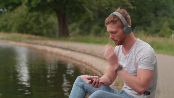 男は池で音楽を聞いて頭をうなずいて土 — ストック動画