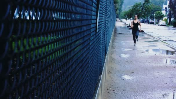 ブルネットの女性はスポーツブラジャーとタイツの砂利道の暗いフェンスに沿って雨の中で実行されます — ストック動画