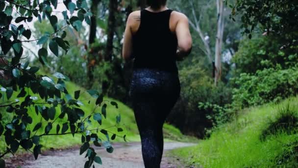 アスレチックブルネットの女性は青い雨の中で緑の森の未舗装の道に沿って逃げる — ストック動画