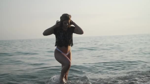 Egy fiatal, szőke, fehér lány fehér bikini alsóban és fekete csipke felsőben kisétál a vízből lassított felvételben..