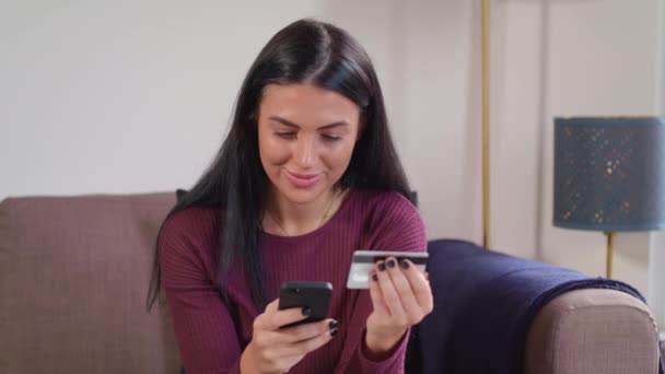 若い女性は オンライン注文をするために彼女の携帯電話を使用してソファに自宅で土 その後 カメラに笑顔 — ストック動画