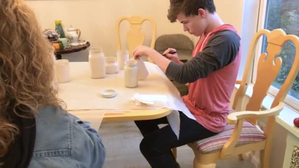 10代の男の子は笑顔で絵付けをしている間に白い絵具でメイソン瓶を花瓶に変える次のイベント — ストック動画