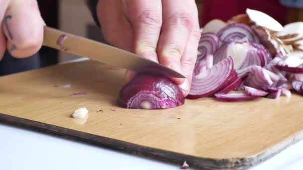 Zblízka na kavkazské mužské ruce v domácí kuchyni. Příprava červené cibule, krájení a nakrájení čerstvých ingrediencí na dřevěné řezací desce.