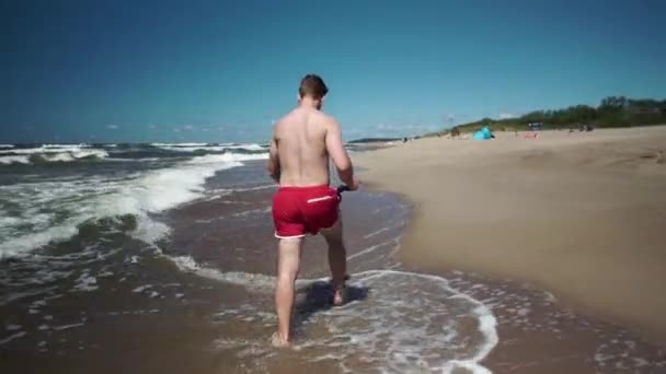 Jovem Calções Vermelhos Caminha Numa Praia Beira Mar Dia Ensolarado — Vídeo de Stock