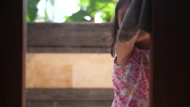 Pretty little Indonesian child helping mom around the house by washing the windows in a door. Child smiles and waves from other side of glass she is wiping clean from the outside.