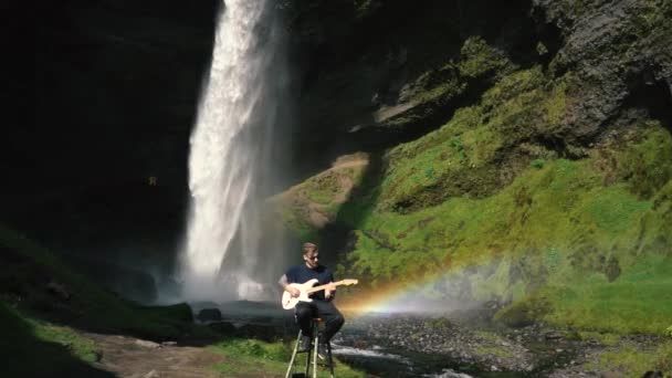 Egy férfi gitározik egy gyönyörű vízesés előtt Izlandon. Lassú Mo és valós idejű felvételek a Sony a7iii és Ronin S-től.