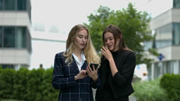 Two business woman looking at a mobile phone in urban environment, discussing over an email.