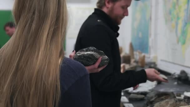 Egy fiatal lány egy nagy követ tart, míg egy férfi egy kisebb követ dob a háttérbe. A diákok elvonják a figyelmüket a főiskolai geológia órán való tanulásról..