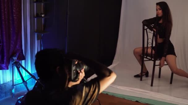 男摄影师拍了几张年轻女模特坐在她身后有白色背景的椅子上的纵向照片 — 图库视频影像
