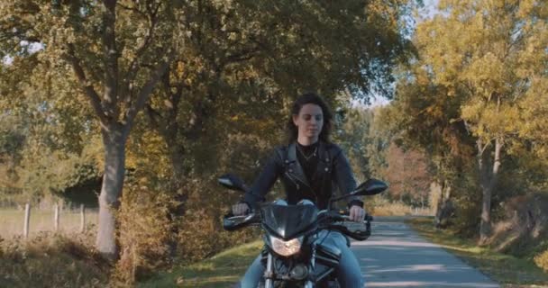 美しい笑顔ヨーロッパ女性モーターバイカー革のジャケット運転を身に着けています秋の紅葉とフィールドに道路のパンニング晴れた日に森の中の木 オランダのユトレヒトでのスローモーション — ストック動画