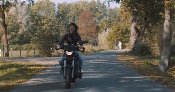 活気に満ちたカラフルな黄金の秋の葉と森の中で革のジャケットを着てバイクを運転かなり笑顔ヨーロッパの若い女性は晴れた日に残します 正面図 スローモーション ユトレヒト オランダ — ストック動画