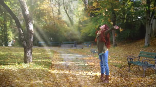 スローモーション 美しい若い赤毛の女性が秋に公園に立っていて 葉が彼女の周りに落ちています 古いベンチと太陽の光で覆われた道を残します ワイドショット スライトパン — ストック動画