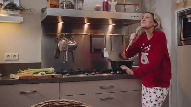 若い女性は食事を準備しながら 彼女のキッチンでお祝いのクリスマスセーターで歌い 音楽を聞いて楽しい — ストック動画
