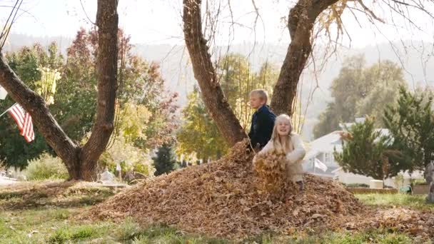 Dvě děti šťastně házejí podzimní listí, zatímco sedí na hromadě. Natočeno v 120fps pomalém pohybu.