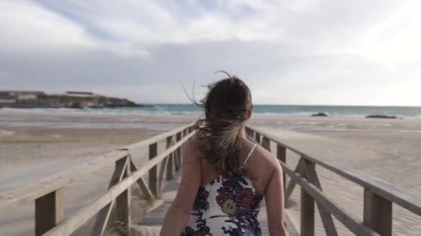 若い女性はカメラから逃げ出し スペインのジブラルタルの砂浜で木製のボードウォークを走っています — ストック動画