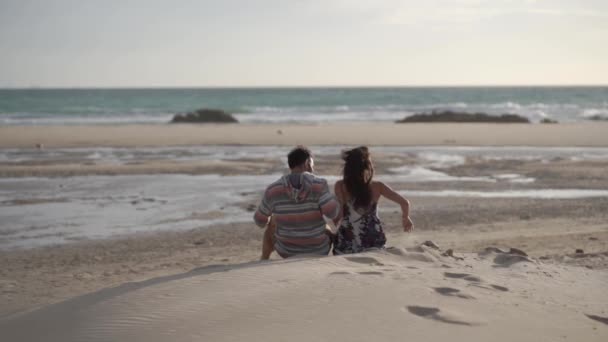 Young couple sit on sandy beach with backs to camera, they embrace and kiss. Slow motion.