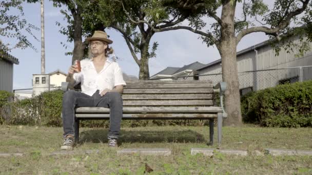 Egy férfi kalapban és szemüvegben ül egy padon a parkban pihentetve és kortyolgatva egy korty sört..