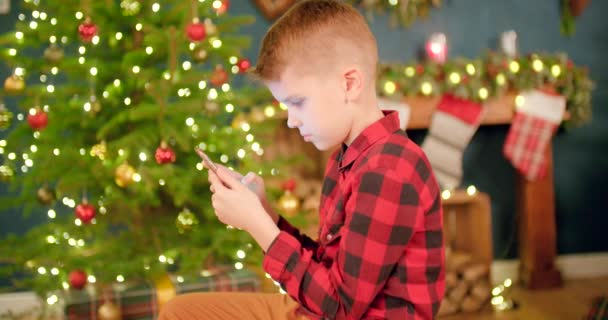 男の子はクリスマスの装飾されたリビングルームで彼の携帯電話で遊ぶことに焦点を当てています — ストック動画