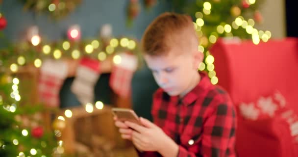 男の子はクリスマスに装飾されたリビングルームに座っている間 スマートフォンを見つめています — ストック動画