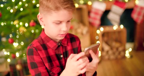 男孩在装饰过的圣诞树前和客厅前的智能手机上打字 — 图库视频影像
