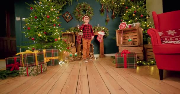 サンタの帽子をかぶっている少年は クリスマスの装飾されたリビングルームの幸福の床の上にスライドしています — ストック動画