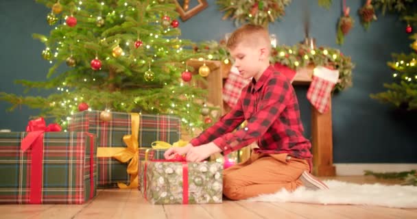 男の子はクリスマスツリーの前で贈り物を包んでいて 中を見ると驚いています — ストック動画