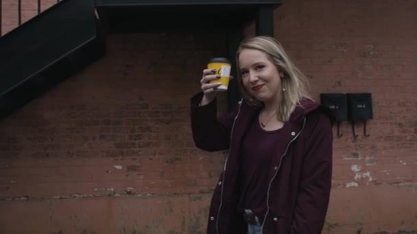 Slow Motion of a Young Woman Smiling and Walking as She Raises Her Coffee Cup Towards the Camera with a Red Brick Wall and Staircase in the Background