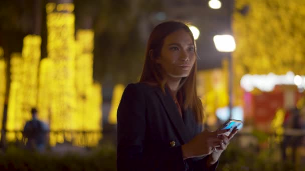 迷人的年轻女子检查了她的智能手机 然后在明亮的城市灯光下对着相机微笑 模糊的背景 — 图库视频影像