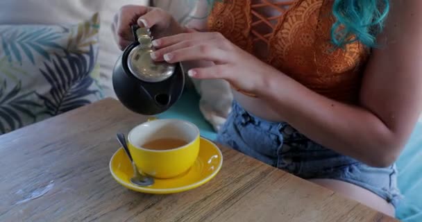 オレンジ作物のトップの青い髪の千年紀の女性は黄色のカップと飲み物にお茶を注ぐ — ストック動画