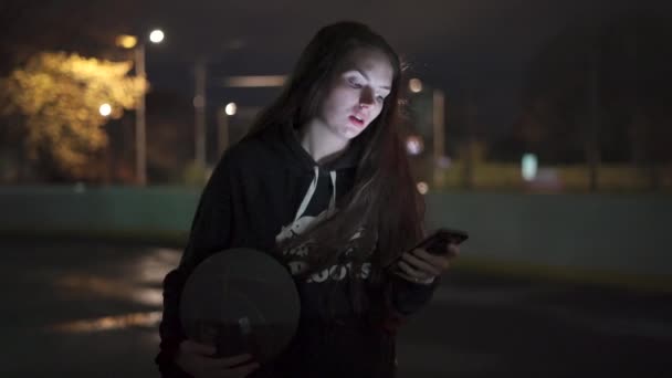 Tiener Meisje Met Behulp Van Telefoon Houden Basketbal Het Buitenterrein — Stockvideo