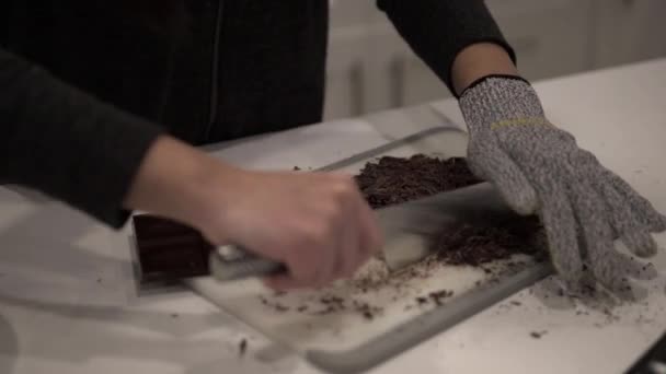 A young girl cuts up dark chocolate before melting it for millionaire shortbread cookies while wearing safety gloves