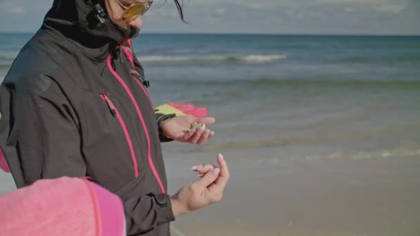 年轻女子戴着太阳镜 头戴头巾 带着小女儿在波罗的海空旷的沙滩上散步 展示了收集到的贝壳和琥珀 慢动作 — 图库视频影像