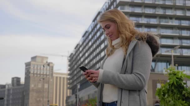 彼女のスマートフォンを使ってかわいい若いブロンドの女性 背景の都市 低角度のパララックスショット — ストック動画