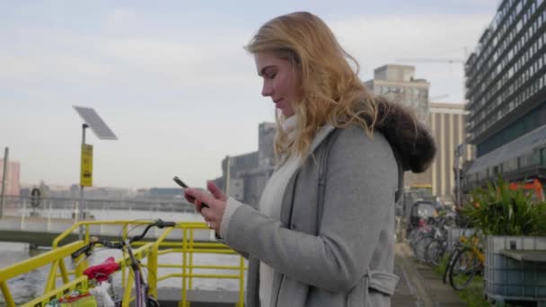 トレンディな若い女性がボードウォークに立って彼女の携帯電話を使用しています 背景にある街 パララックスショット — ストック動画