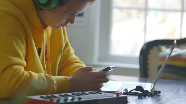 テーブルの上にミディキーボードでノートパソコンに座っている間 Iphoneとヘッドフォンの十代の男の子 — ストック動画