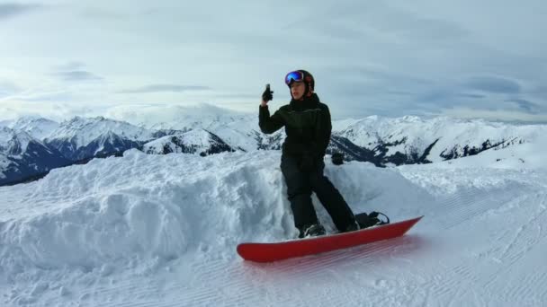 Tinédzser snowboardos szelfit készít a havon ülve, hegyekkel körülvéve..