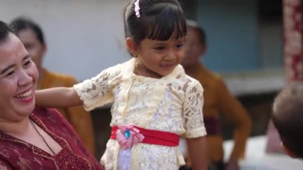 穿着白花边衣服的可爱小女孩弯腰吻着婴儿的脸颊 拍拍他的头 在巴厘岛一座寺庙举行的印度教仪式上 妈妈抱着年幼的女儿向朋友们打招呼 — 图库视频影像