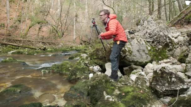 Άνθρωπος Στήνει Ένα Τρίποδο Δίπλα Ένα Ποτάμι Λαμπερό Πορτοκαλί Μπουφάν — Αρχείο Βίντεο