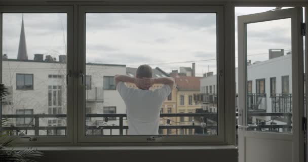Široký statický záběr kamer norského muže izolovaného v karanténě viru korony, jak se natahuje dozadu a dívá se na lidi z balkónu, natáčené v tmavém bytě, v Oslo Norsko.